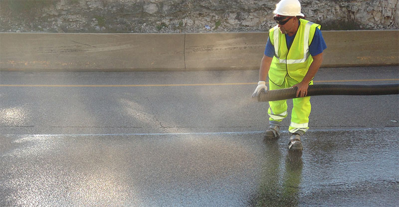 Photo of worker applying aggregate over polymer on asphalt pavement.
